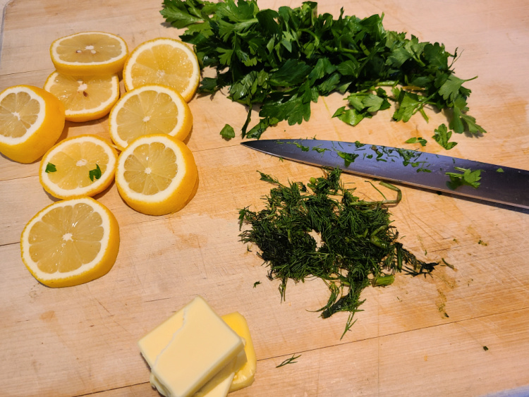 Cut butter slices, lemon slices, and fresh minced parsley and dill on a cutting board with a knife 