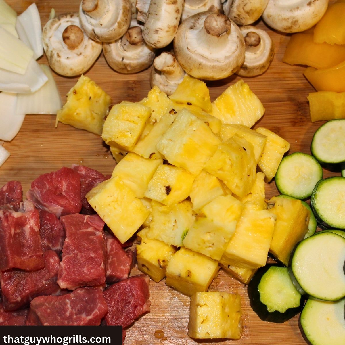 Cubed Steak on a cutting board with cut pineapple, sliced zucchini, mushrooms, yellow pepper, and white onions