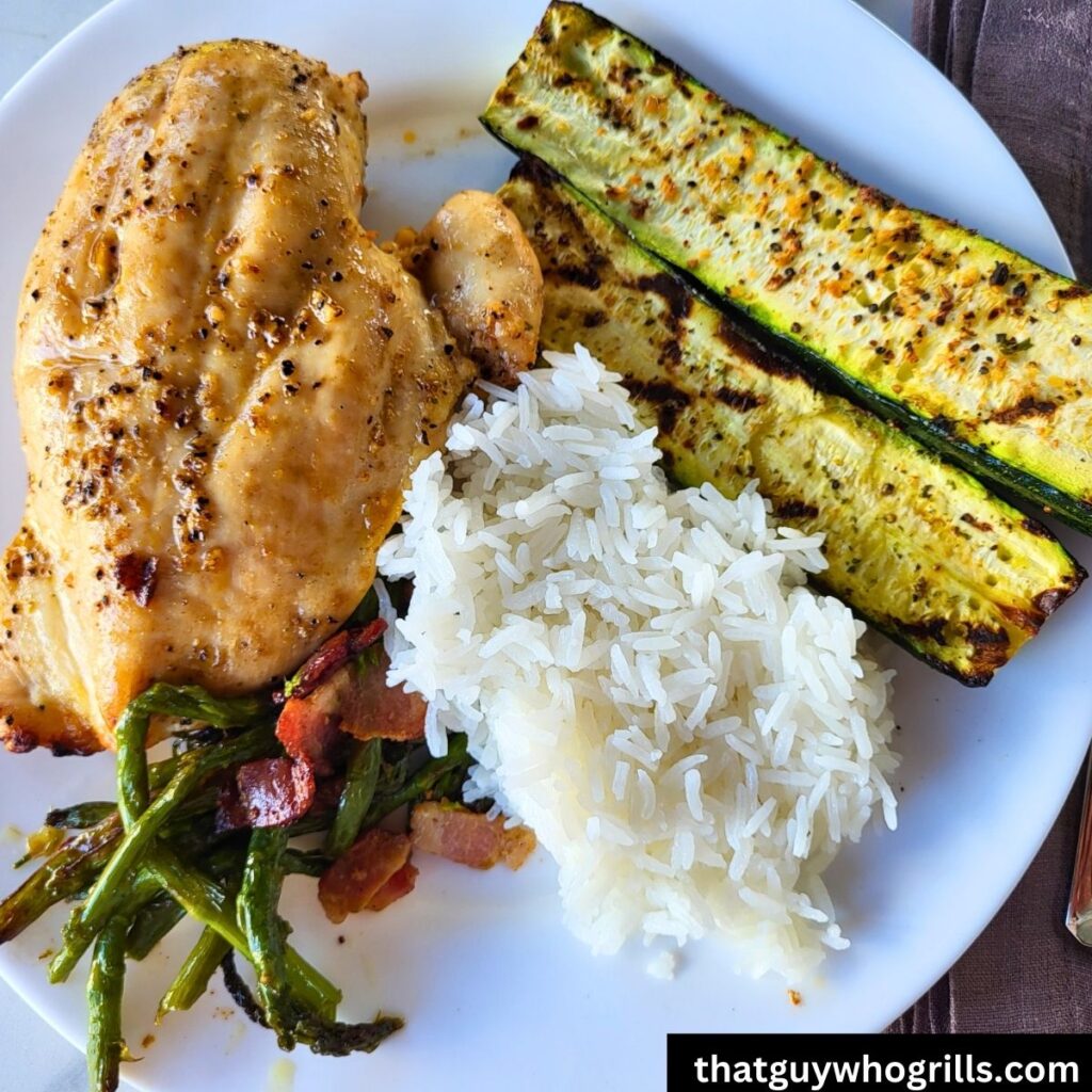 Grilled Chicken breast with white rice, bacon and asparagus, and grilled zucchini on a white plate